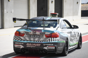 Rear view of a BMW M4 race car with a large carbon-fiber wing, aggressive diffuser, and sponsor decals, parked in a pit lane.